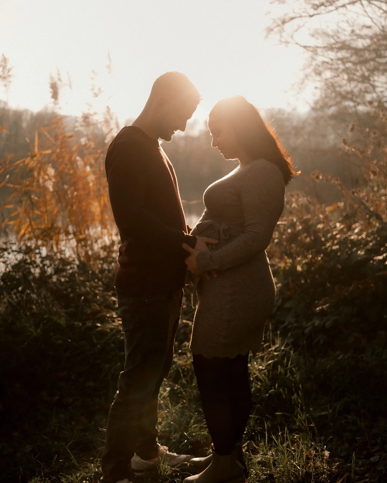 Werdende Eltern stehen sich im Herbstlicht gegenüber, seine Hände auf ihrem Babybauch