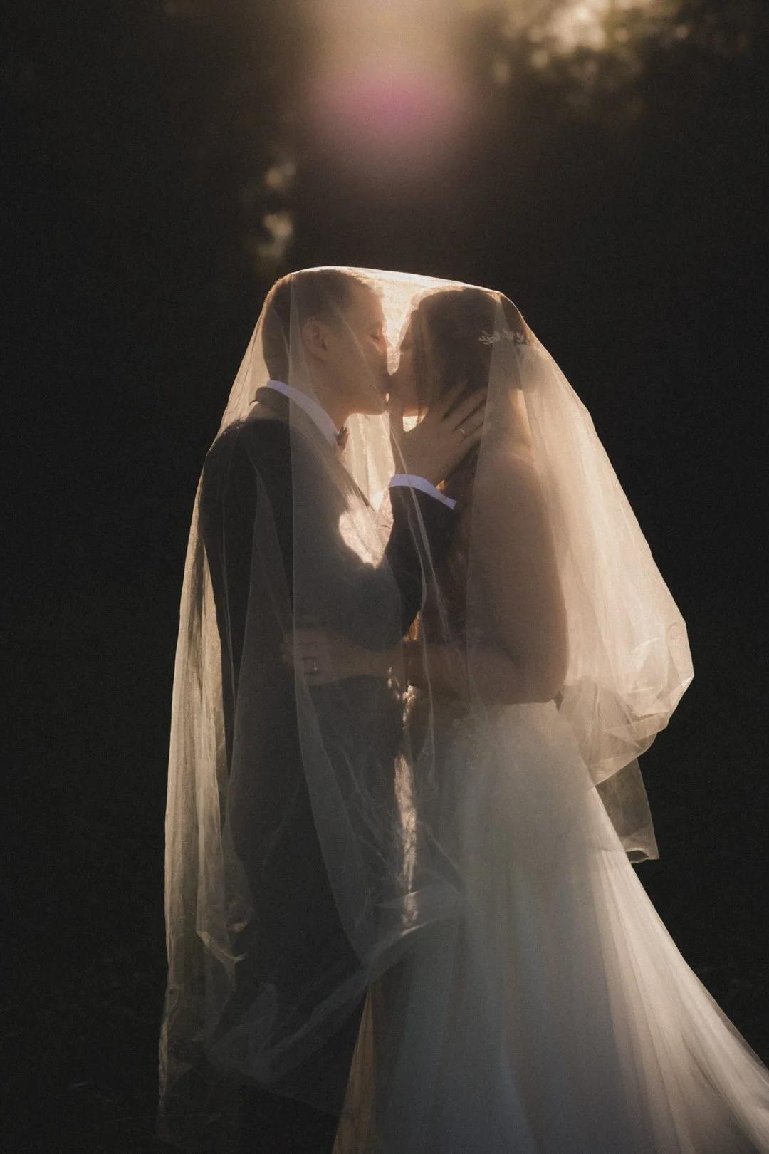 Brautpaar küsst sich unter dem Schleier im warmen Gegenlicht – Hochzeitsfotografie
